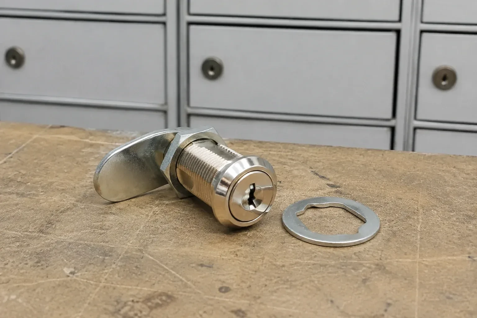 Detached threaded cam lock and mounting washer on a workbench in front of closed mail compartments. — Mailbox Lock Replacement, Oakville
